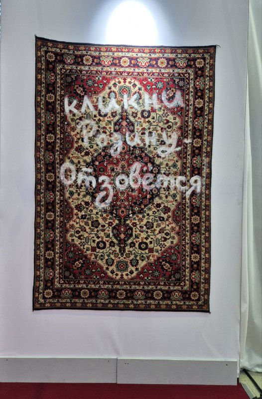 Гобелен или ковёр, подвешенный на стене, с надписью поверх орнамента — инсталляция с традиционной текстильной фактурой и современным посылом.