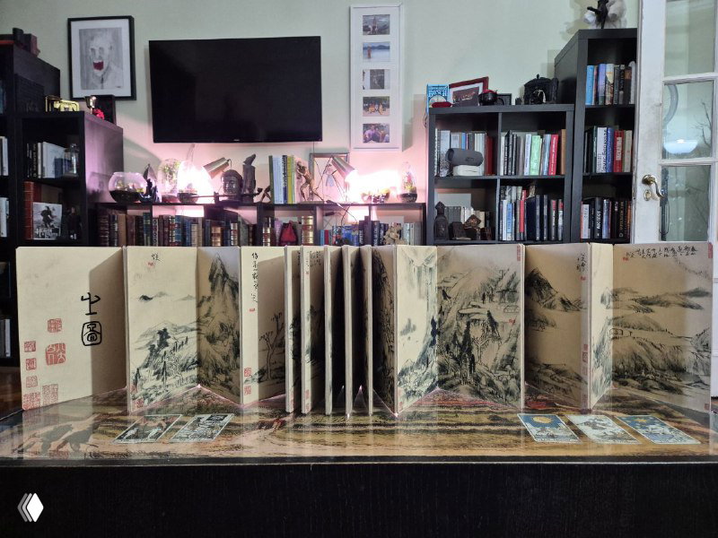 Раскладная панорама книги в развёрнутом виде на столе: ряд миниатюрных листов с тушевыми китайскими пейзажами и каллиграфией на заднем плане книжных полок.