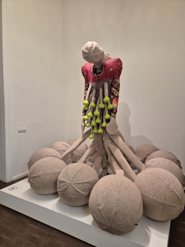 Скульптурная работа в экспозиции: текстильные шарообразные объекты и центральная фигура с яркими элементами на фоне белых стен зала.