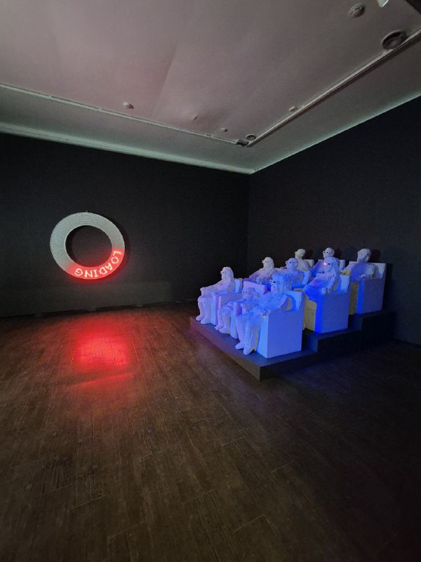 Инсталляция с неоновым кольцом на стене и белыми скульптурными фигурами, расставленными в полутемном зале музея.