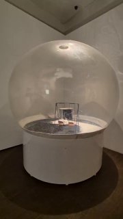 Инсталляция под прозрачным куполом: круглый подиум и внутри небольшие объекты, отсечённые светом в затемнённом пространстве зала.