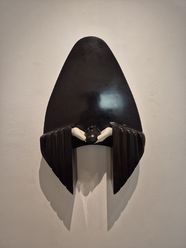 Тёмная скульптурная форма с белыми элементами в центре, напоминающая маску или щит, расположенная на стене экспозиционного зала.