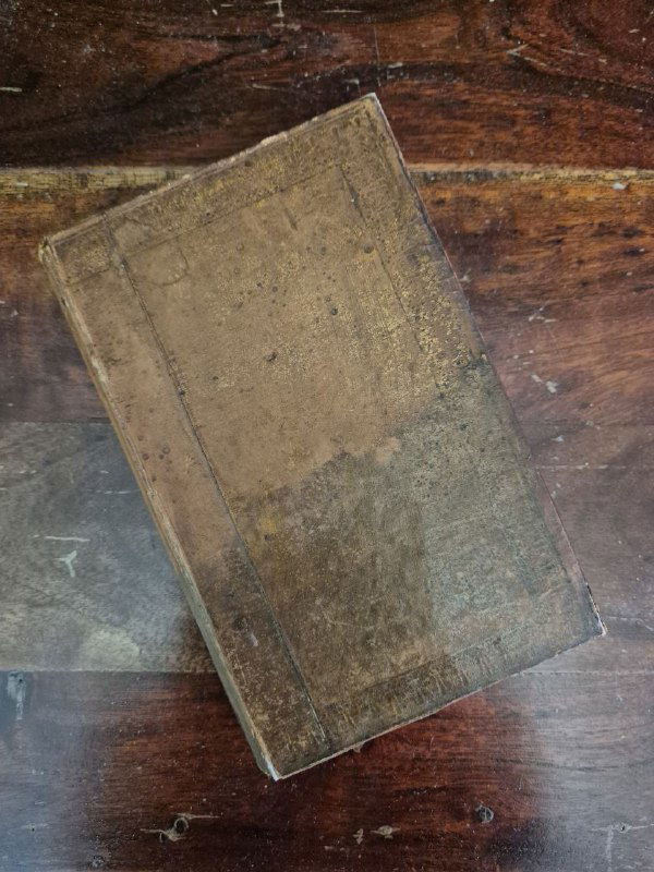 Вид сверху на старую книгу в коричневом переплёте, лежащую на столе; заметны пятна и следы времени, подчёркивающие подлинность издания.