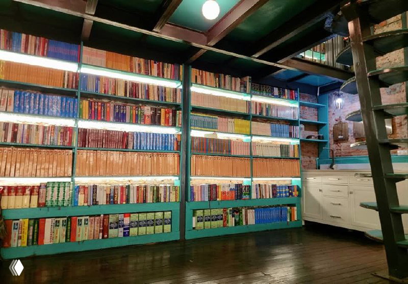 Книжные полки, плотно уставленные разноцветными корешками, тёплое освещение и часть мебели в книжном лаундже гостевого дома.