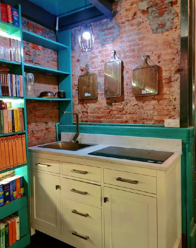 Небольшая кухонная зона внутри гостевого дома: раковина, шкафчики и открытые полки рядом с кирпичной стеной и книгами на стеллажах.