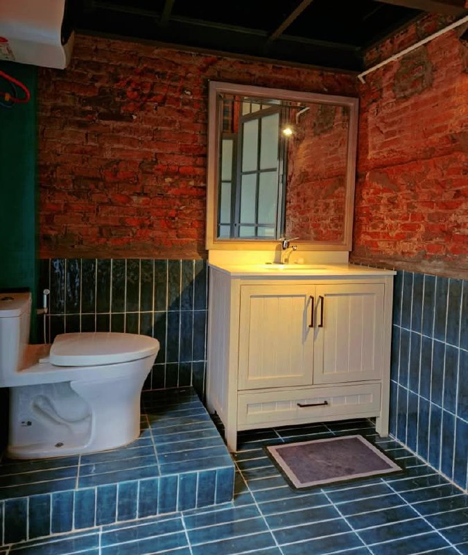 Ванная комната гостевого дома: унитаз и тумба с раковиной на фоне оголённой кирпичной стены и плиточного пола в зелёных тонах.
