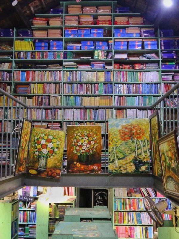 Высокие полки до потолка с рядами книг и картинами на переднем плане — многоуровневый книжный интерьер в Nhà sách Mão.