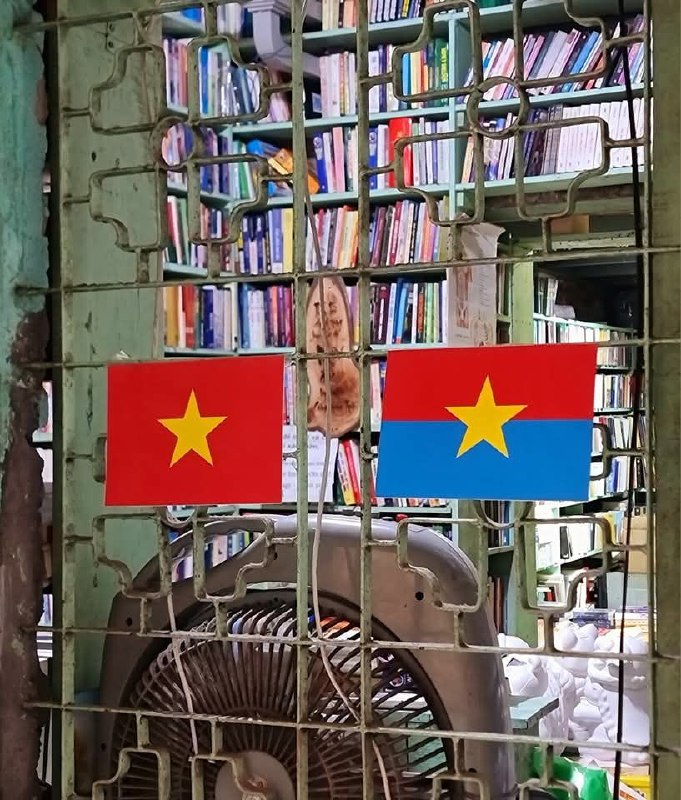 Крупный план решётки с двумя флажками и задним планом из книг на полках — декоративный элемент внутри магазина Nhà sách Mão.