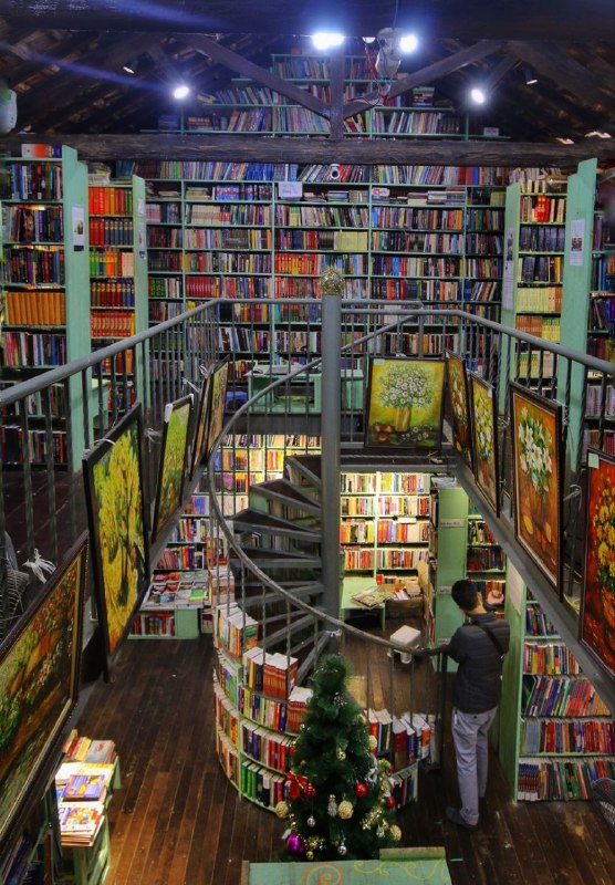 Общий вид большого двухуровневого книжного пространства Nhà sách Mão с винтовой лестницей, центральным проходом и посетителем у полок.