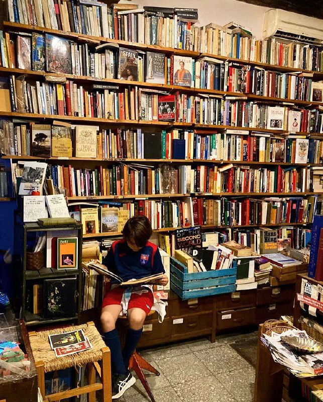 Ребёнок, читающий книгу у низких стеллажей в магазине: коробки с изданиями, детский стул и домашняя, дружелюбная обстановка, характерная для букинистики.