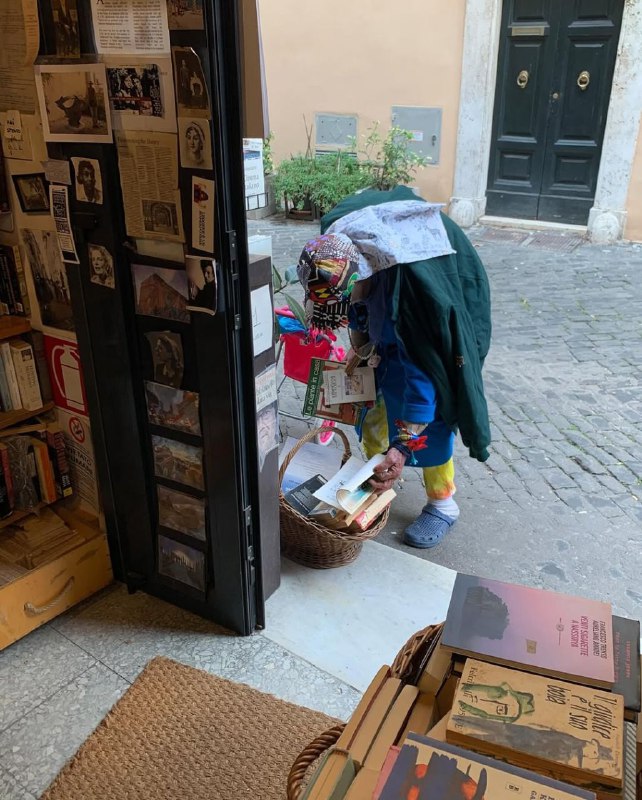 Сцена у порога: посетитель наклоняется над корзиной с книгами, видно открытый вход на мощёную римскую улицу и соседний тихий дворик Трастевере.