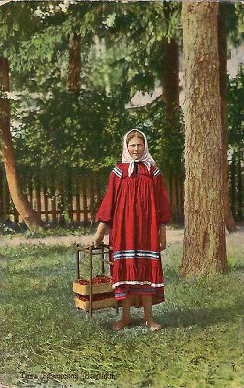 Женщина в красном платье с коробками в руках, стоит у деревянного забора в парковой зоне; цветная почтовая открытка начала XX века.