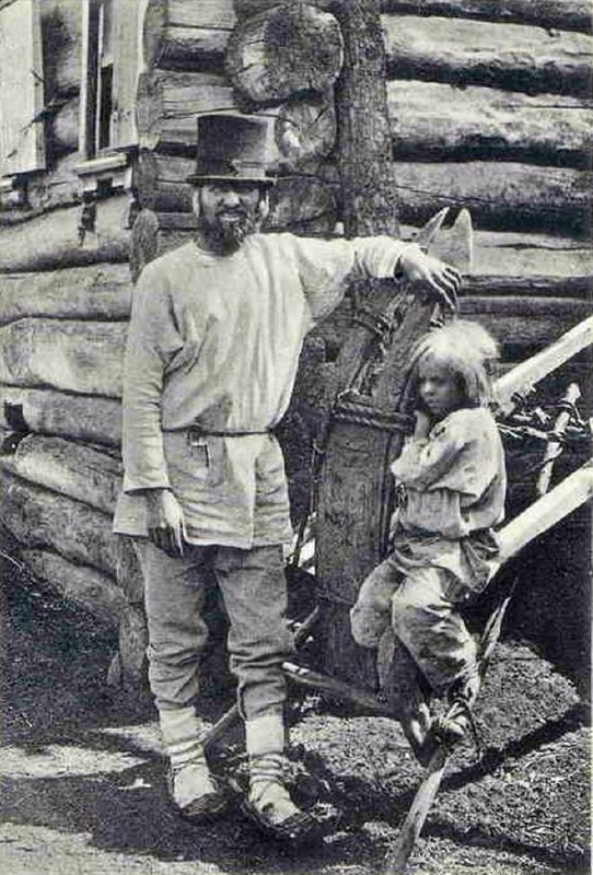 Мужчина в шляпе с ребёнком у сруба дома; чёрно‑белая почтовая фотография с типичным сельским портретом начала XX века.