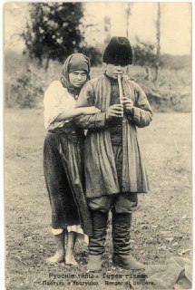 Юноша с деревянной флейтой и девушка рядом в национальных костюмах; монохромная композиция с народными мотивами начала XX века.