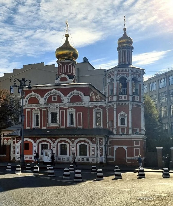 Современное фото Храма Всех Святых на Кулишках: красно‑белые стены, купол с крестом и городской фон из окружающих построек при ясной погоде.
