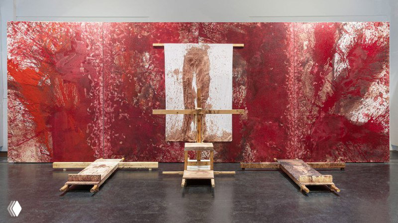 Широкая инсталляция: центральная фигура на фоне большого полотна, облитого бордовыми и красными пятнами, на полу видно продолжение рисунка краски.