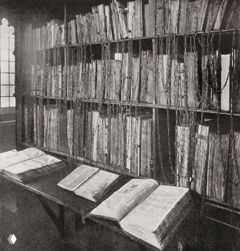 Чёрно‑белая историческая фотография полки с прикованными цепями книгами: видны корешки томов, металлические цепи и окно библиотеки позади.
