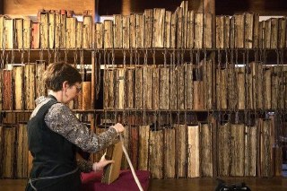 Сотрудница библиотеки у стеллажа с прикованными книгами: она измеряет или перебирает том, видны тонкие цепочки и старые переплёты.