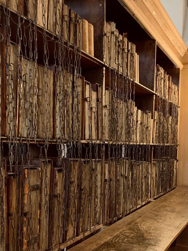 Низкий ракурс полки с прикованными книгами: длинные цепи, упорядоченные тома и тёмное дерево полок создают атмосферу старинного архива.