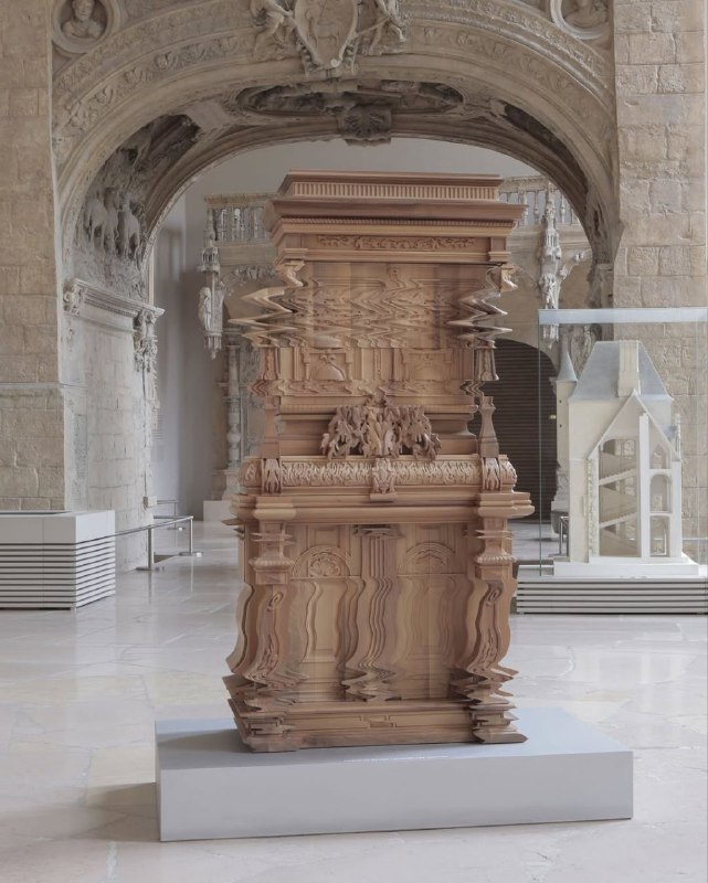 Скульптура в музейном пространстве: деревянный монумент на постаменте, изображение которого кажется частично «съёмочным» и деформированным как цифровой сдвиг.