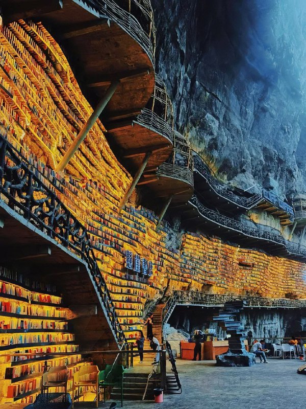 Интерьер пещерного книжного магазина: тёплое освещение, ступени и полки с книгами, уютная зона чтения у подножия стены.