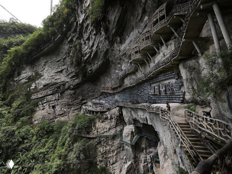 Наружная тропинка, обвивающая скалу между растительностью и вертикальными полками; вид на фасад книжного магазина со стороны.