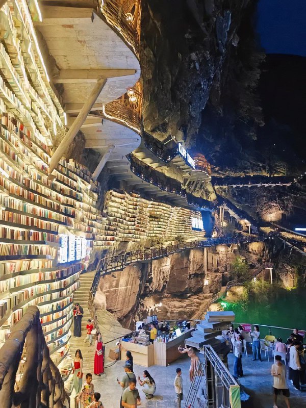 Ночная сцена: подсвеченные полки и посетители у нижней площадки пещерного книжного магазина.