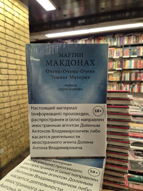 Обложка сборника Мартина Макдонаха «Очень-очень-очень темная материя» в магазине; на обложке видна наклейка с возрастным ограничением и стопки книг вокруг.