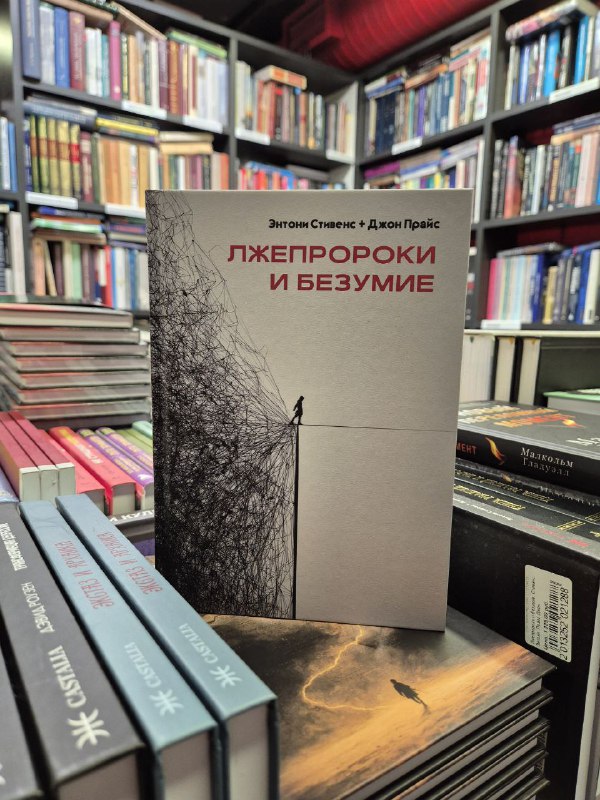 Книга «Лжепророки и безумие» Стивенс и Прайс на полке книжной лавки: минималистичная обложка выделяется на фоне других изданий.