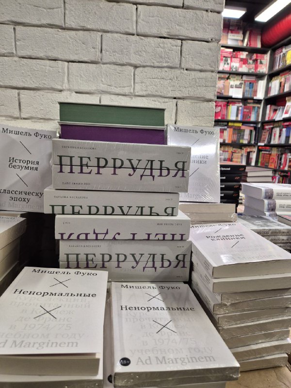 Стопки книг Перрудью в упаковке на прилавке книжного магазина, рядом видны другие издания и кирпичная стена магазина.