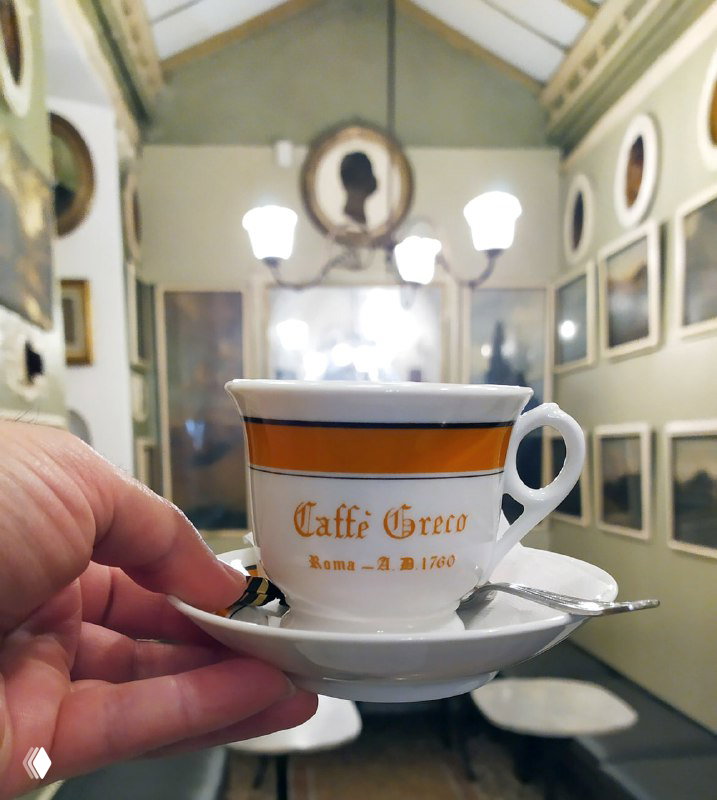 Ручка чашки с логотипом Caffè Greco на фоне зала с портретами: крупный план чашки, мраморные столики и декор кафе видны позади.
