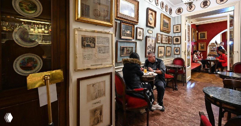 Уютный зал кафе Caffè Greco: столики, кресла и стены, уставленные картинами и рамами, посетители за беседой в историческом интерьере.