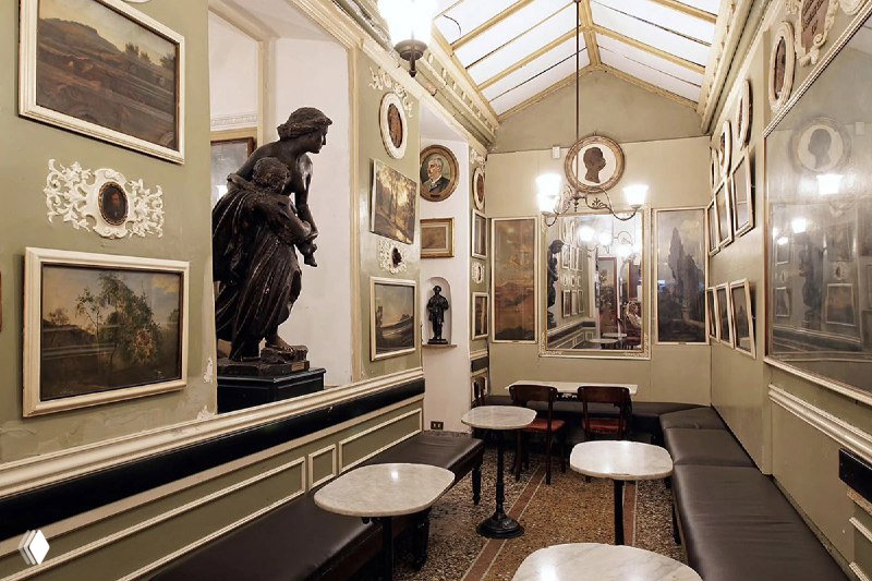 Узкий зал с высокими потолками и галереей портретов по периметру, пустые столики и мягкое освещение — атмосфера старинного римского кафе.