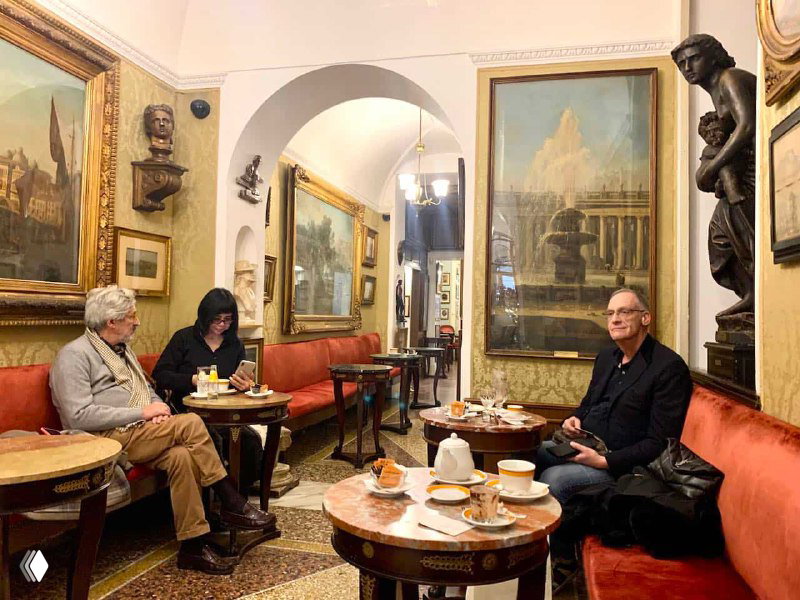 Группа пожилых гостей за столиками в историческом интерьере Caffè Greco: беседа, чашки кофе и картины на стенах вокруг них.