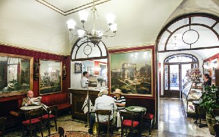 Ещё один ракурс зала Caffè Greco: столики, посетители и большая арочная входная группа, через которую видно внутреннее пространство кафе.