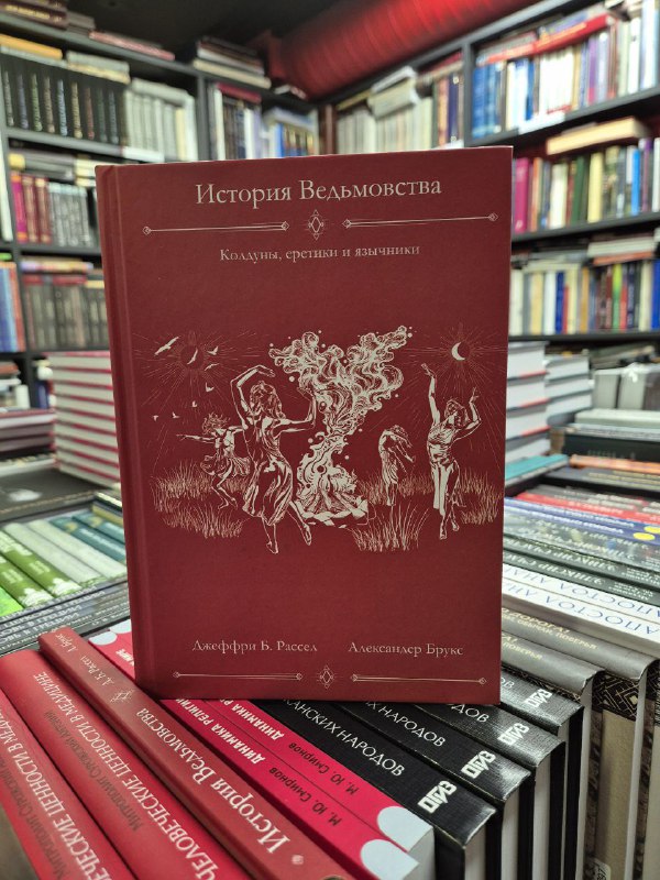 Красная обложка книги «История ведьмовства» Рассела и Брукса в крупном плане на прилавке, видны другие тома вокруг.