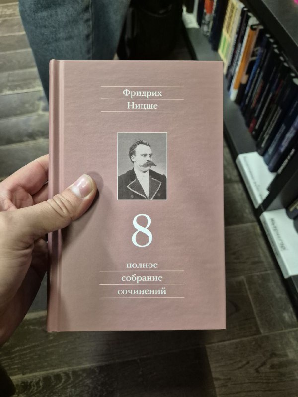 Розовый том «Полное собрание сочинений Фридриха Ницше» в руках читателя на фоне книжных полок, демонстрирующий формат издания.