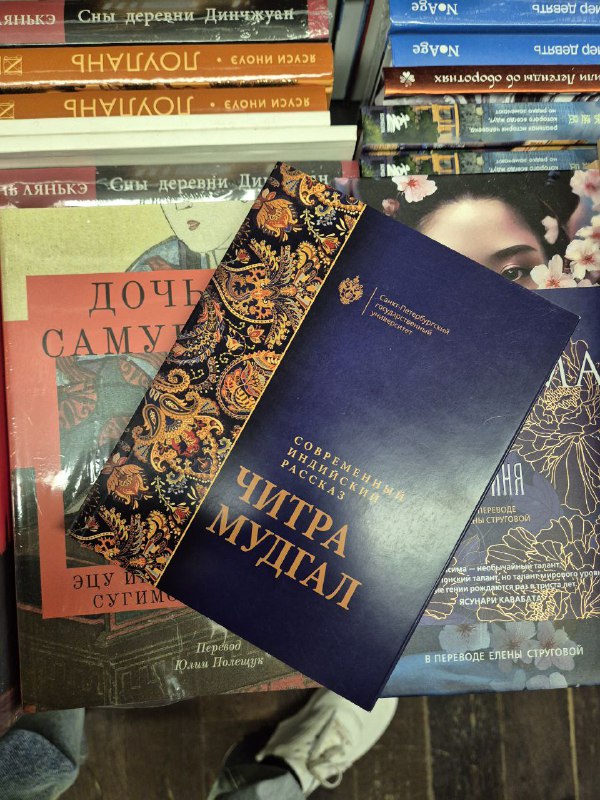 Сборник современных индийских рассказов Читры Мудгал с темно-синей обложкой и золотым тиснением, лежащий на стопке прочих новинок.