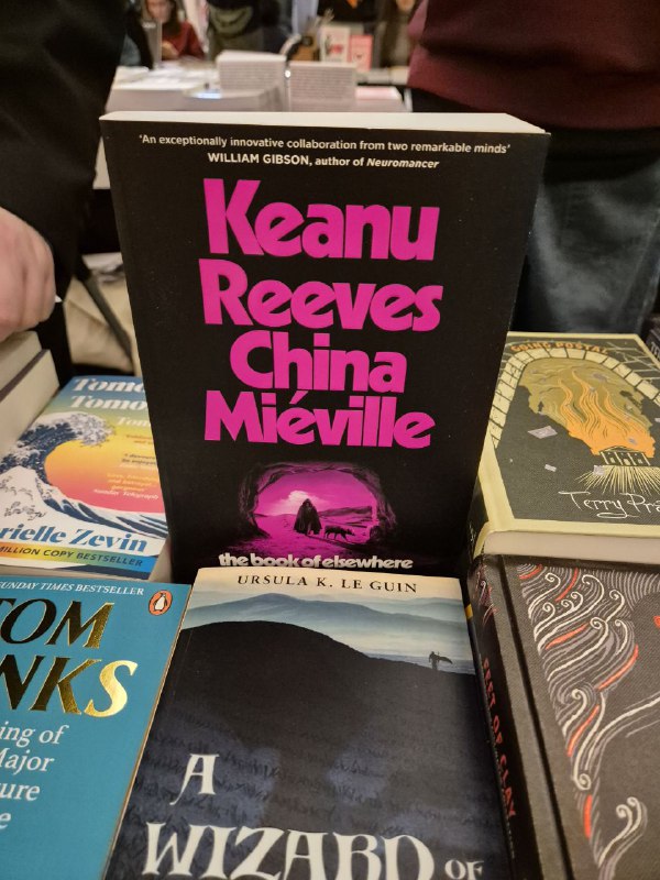 Книга с фиолетовой обложкой 'Keanu Reeves China Mieville' на столе ярмарки Rassvet, вокруг видны другие книги и бумажные ценники.
