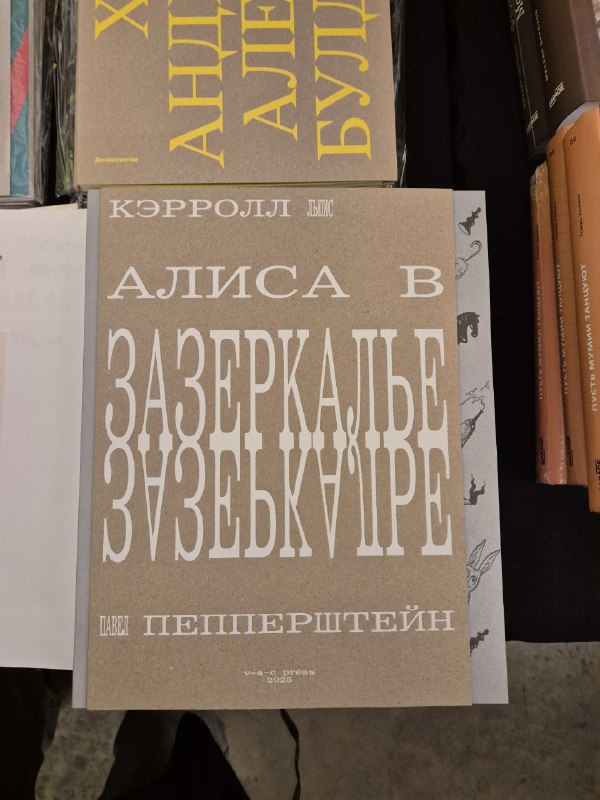 Серо-бежевый переплет книги 'Алиса в Зазеркалье' на прилавке ярмарки Rassvet, видно типографику и соседние издания рядом.