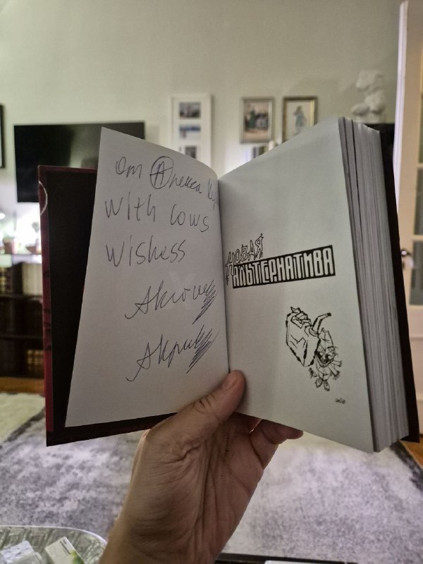 Открытая книга показывает посвящение и подпись на внутренней странице; снимок сделан в комнате с книжными полками и мебелью на заднем плане.