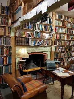 Интерьер книжного магазина: плотно уставленные стеллажи с подержанными книгами, уютное кресло, письменный стол с печатной машинкой и настольной лампой.