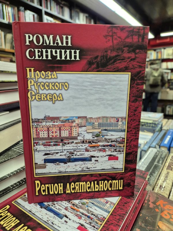 Книга Романа Сенчина «Проза Русского Севера» в красной обложке на столе книжного магазина, вокруг видны другие экземпляры и полки.