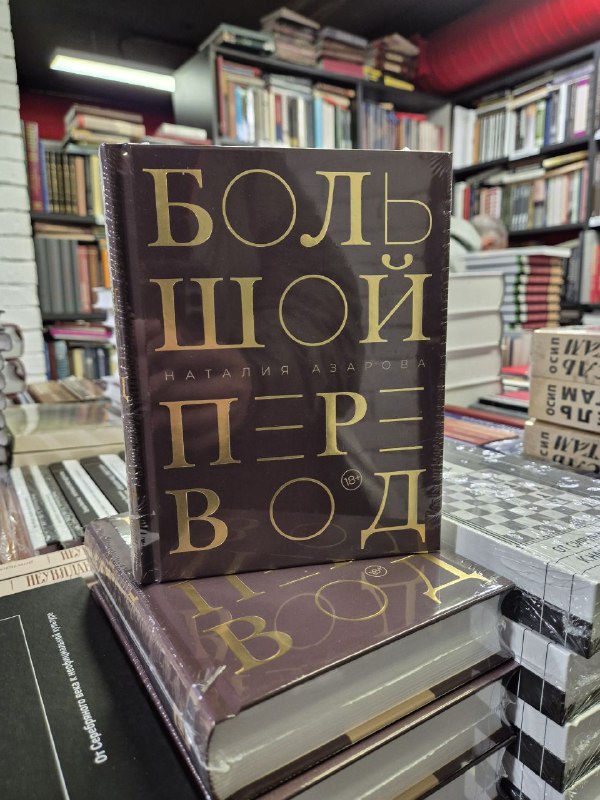 Крупный план книги «Большой перевод» Наталии Азаровой в плотной обложке на столе, вокруг стопки других книг в книжном магазине.