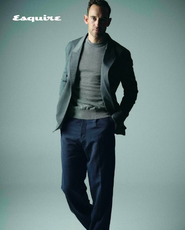 Полнотелая фотография Жоэля Диккера в деловом образе: костюм и свитер, расслабленная поза на однотонном фоне в спокойной цветовой гамме.