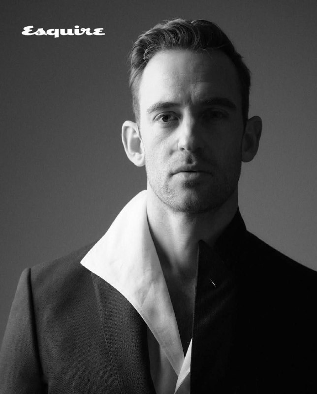 Чёрно‑белый портрет Жоэля Диккера в формальной одежде с приподнятым воротником, строгая композиция и контраст света и тени.