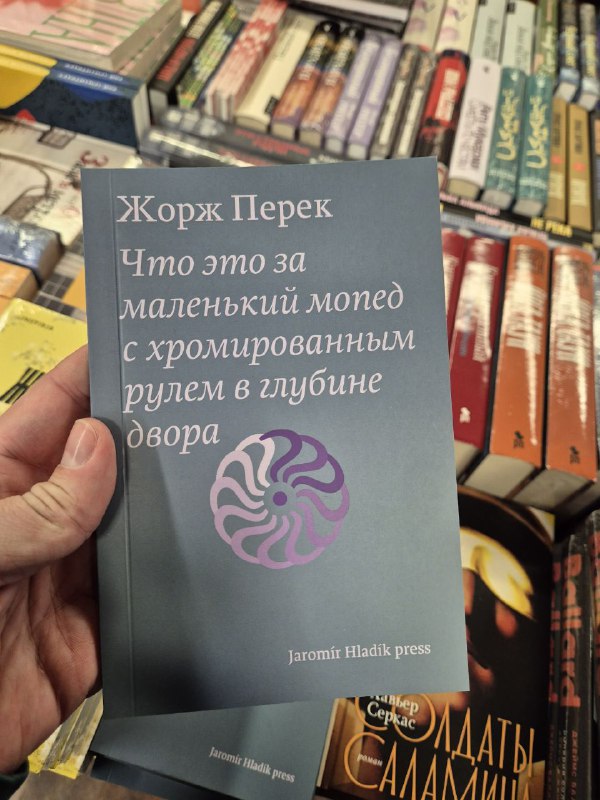 Обложка Жоржа Перека «Что это за маленький мопед с хромированным рулем в глубине двора» на фоне книжной полки, мягкий тон