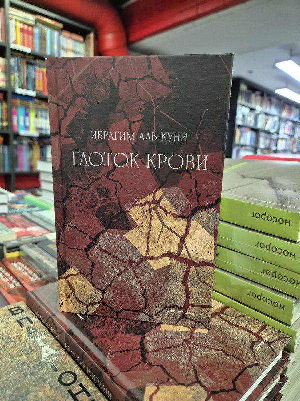 Книга «Глоток крови» Ибрагима аль-Куни с декоративной обложкой, стопка вокруг, книжный магазин в фоне