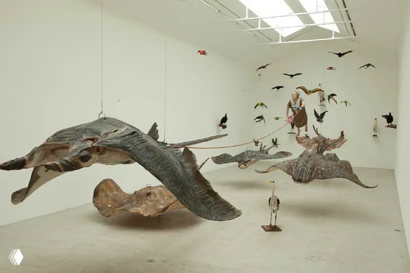 Подвесные скульптуры птиц и фантастических существ в белом выставочном зале; динамичная инсталляция с элементами природы и ремесла.