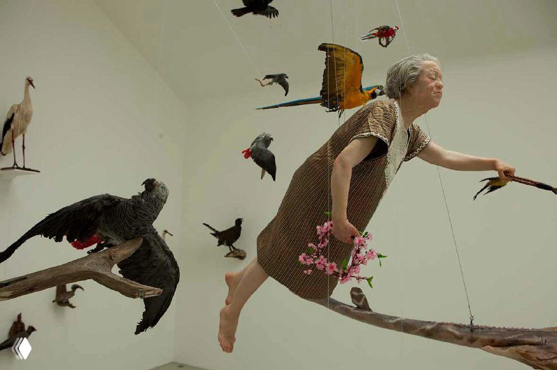 Фигура пожилой женщины, словно летящая среди множества птиц; сюрреалистическая сценография, подчёркивающая темы старения и свободы.
